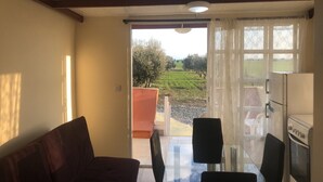 A room with a sofa, a dining table with chairs, a kitchen area with a refrigerator and oven, and a view of a green landscape through a glass door. - MERSINERA Paradise Garden NOAH  house (Trachoni)