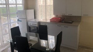 A compact kitchen with white cabinets, a gas stove, a refrigerator, and a sink. There is a dining table with chairs and a window with curtains. - MERSINERA Paradise Garden NOAH  house (Trachoni)