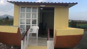 A small yellow building with a red roof, a white door, and a small set of stairs leading to a covered area with a table and chairs. - MERSINERA Paradise Garden NOAH  house (Trachoni)