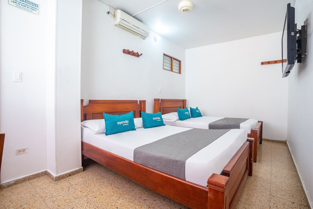 A hotel room with two wooden beds, each with a grey and blue pillow, and a grey blanket. There is a TV mounted on the wall, and an air conditioning unit above the beds.