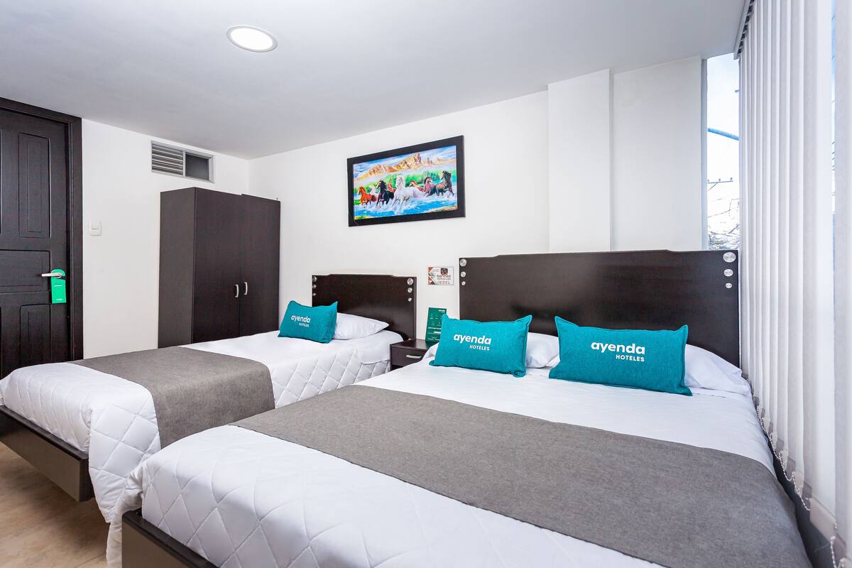 Ein Hotelzimmer mit zwei Betten, jeweils mit einer grau-weißen Daunendecke und türkisfarbenen Kissen mit dem Branding 'ayenda hoteles'.
