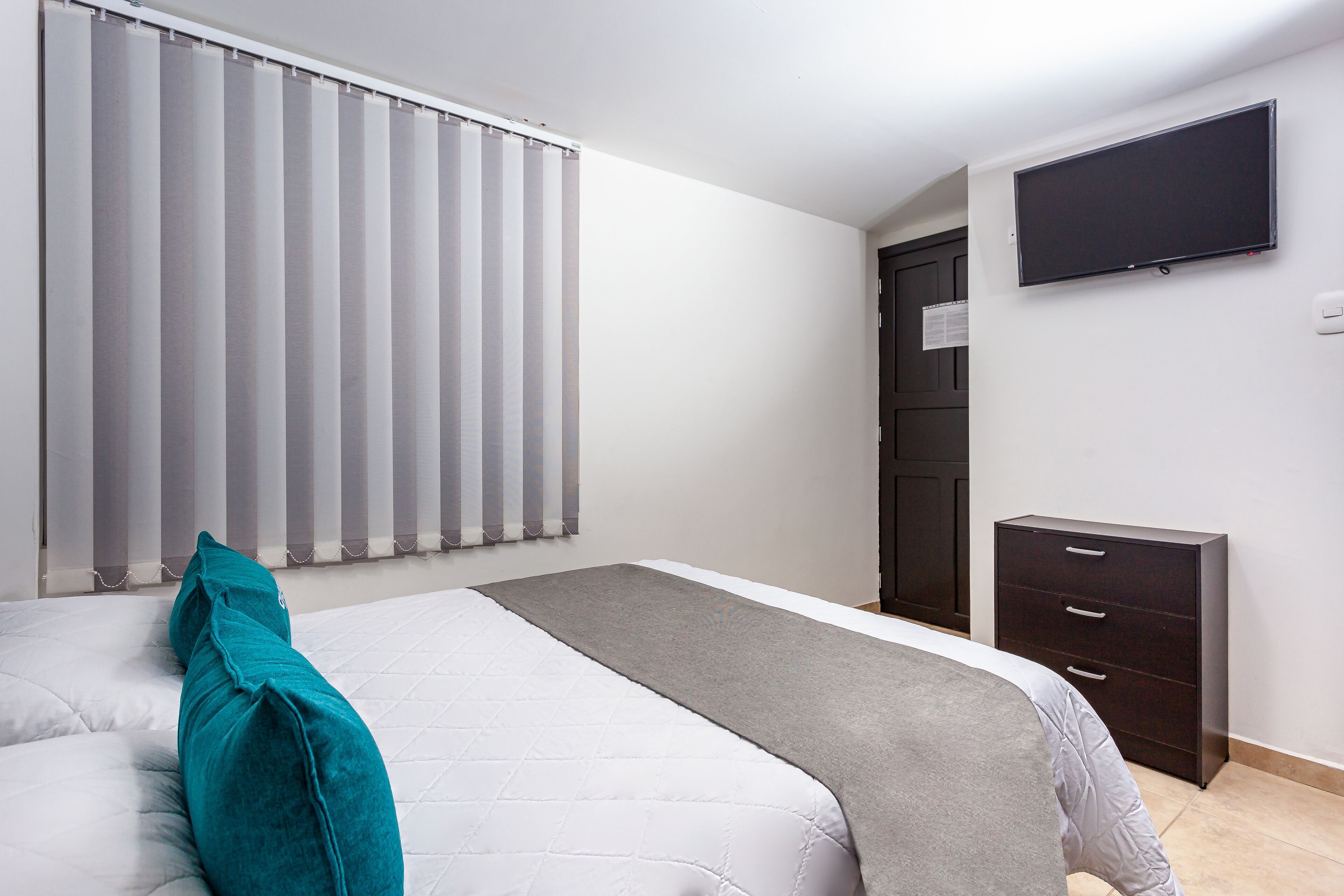 A bedroom with a bed, a TV mounted on the wall, a dark wooden dresser, and a window with blinds.