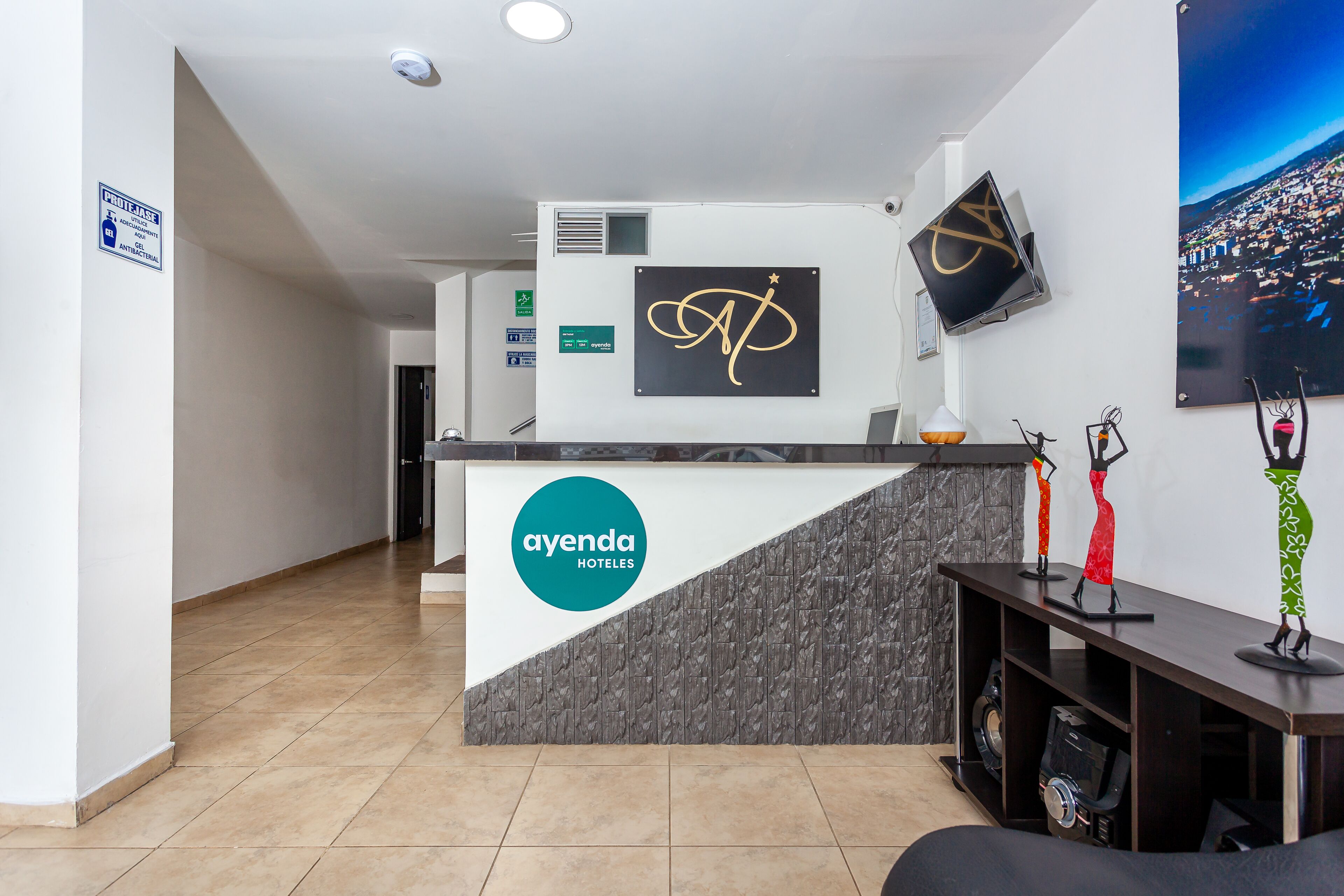 A hotel reception area with a modern design, featuring a reception desk with the logo 'ayenda HOTELES', a mounted television, and a cityscape poster on the wall.