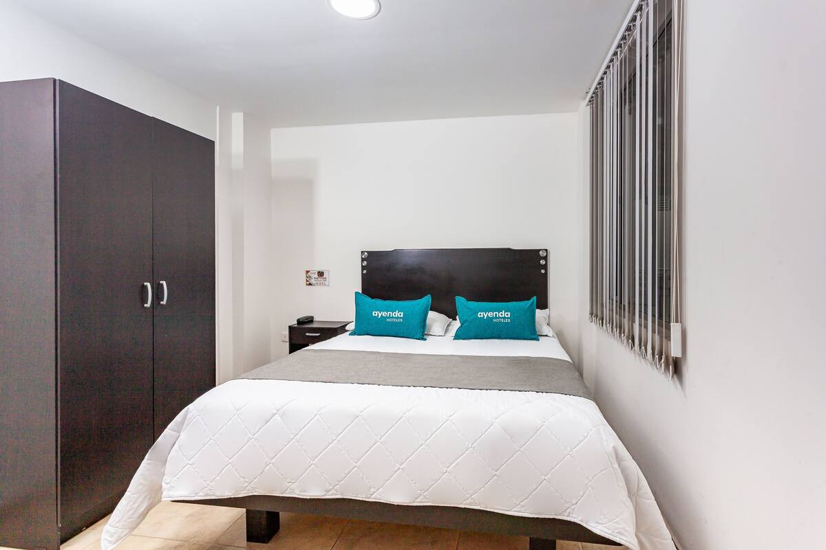 Ein Hotelzimmer mit großem Kleiderschrank, einem Bett mit weißer gesteppter Bettdecke und zwei Kissen mit 'ayenda hotels' Branding, einem Nachttisch und einem Fenster mit Jalousien.