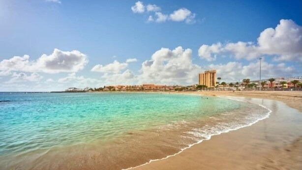 A beach with clear turquoise water, a sandy shore, and a hotel with multiple floors in the background. - Centrally located on the beachfront in Los Cristianos (Arona)
