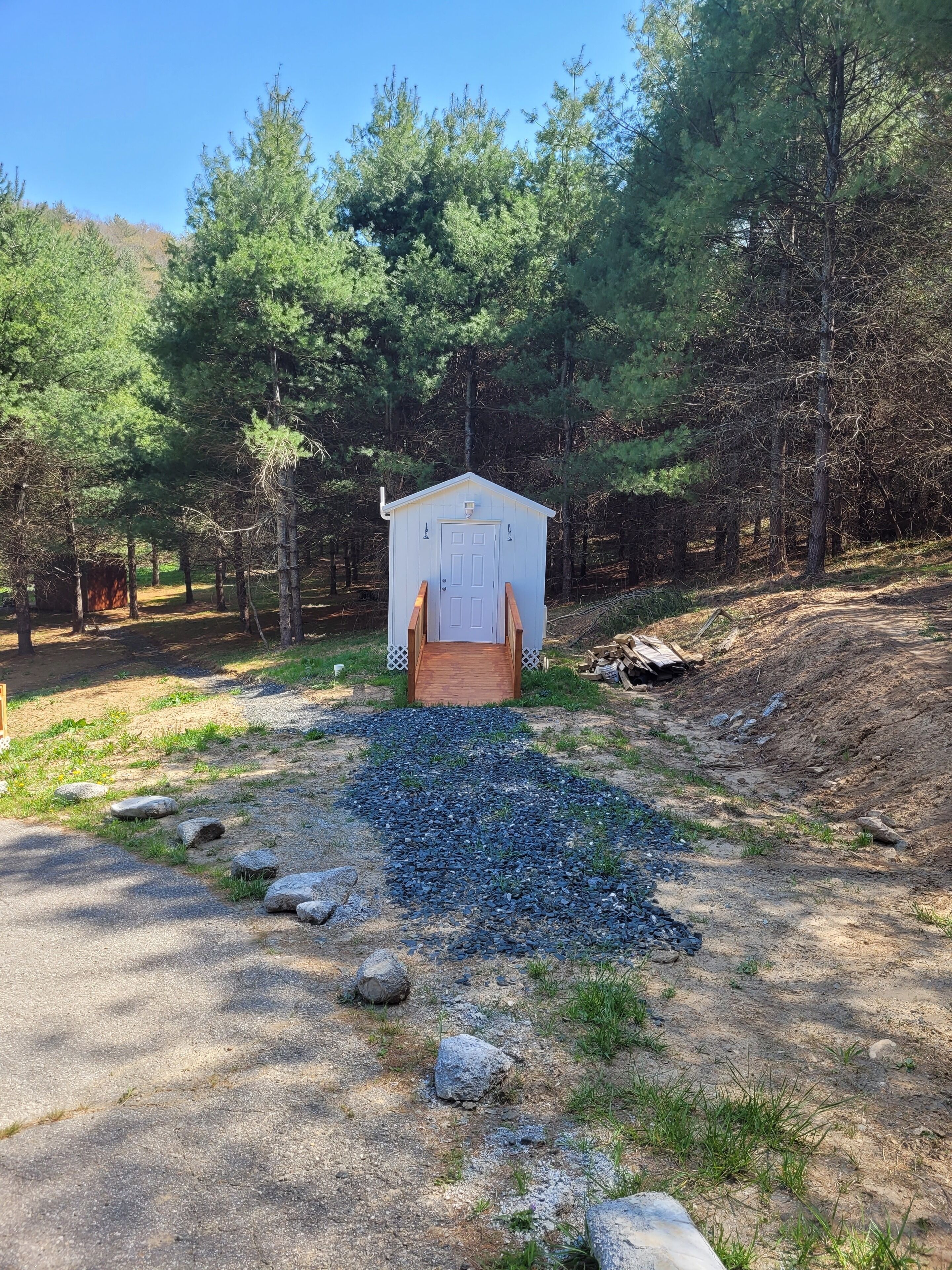A small wooden cabin with a door and windows, surrounded by trees and a gravel path leading to it.