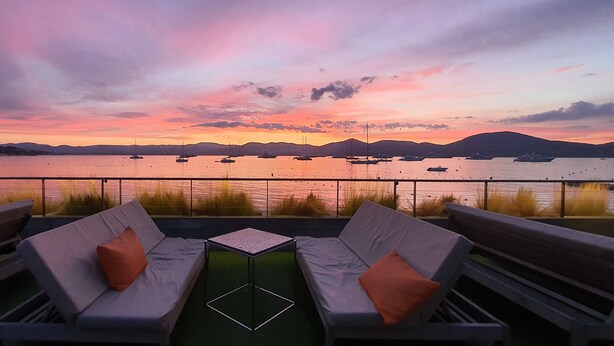 Terraza con dos sofás y una pequeña mesa, con vista a una masa de agua tranquila con barcos y colinas lejanas.