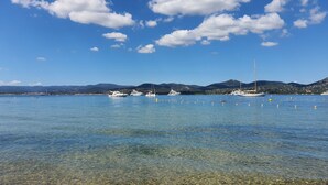 Vista despejada de una zona costera con barcos anclados en el agua, una playa arenosa y montañas lejanas bajo un cielo azul con nubes.