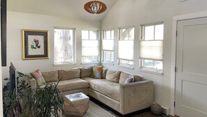 Una sala de estar con un sofá seccional color beige, una mesita de centro pequeña y una planta en maceta. Hay varias ventanas con persianas, una puerta y un cuadro en la pared.