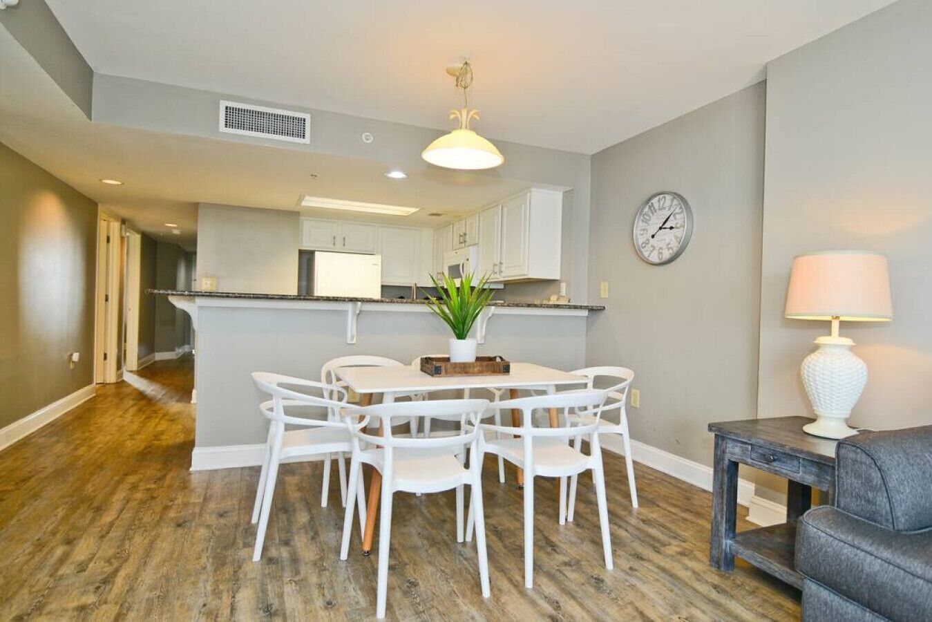 A modern kitchen with white cabinets, a dining area with a round table and white chairs, a clock on the wall, and a lamp on a side table.