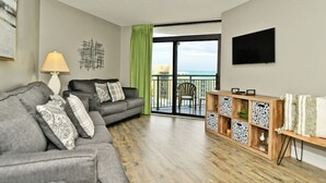 Ein modernes Wohnzimmer mit grauem Sofa, einem Holztisch und Blick auf das Meer vom Balkon.