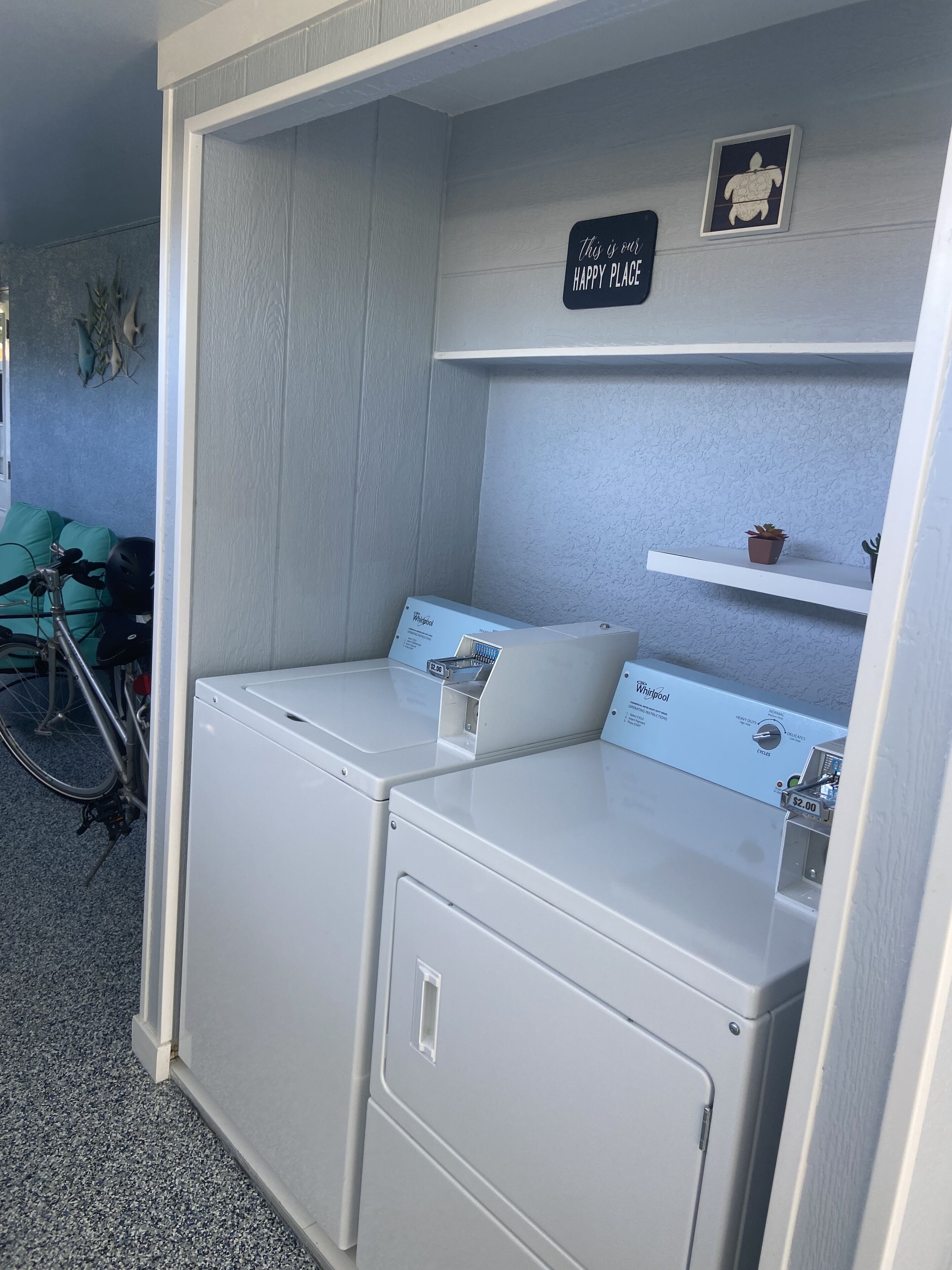 Uma lavanderia com duas secadoras brancas Whirlpool, uma prateleira com a placa 'Happy Place' e uma bicicleta ao fundo.