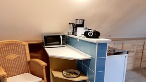 Een kleine keuken met magnetron, koffiezetapparaat en een kleine koelkast. Er staat een rieten stoel met kussen en een plank met servies.