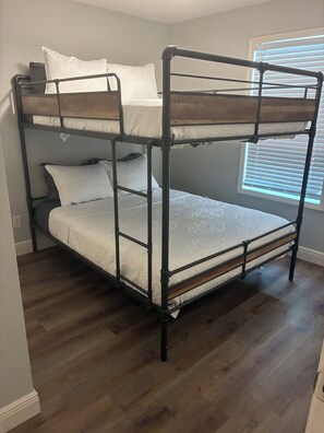 Een stapelbed met een houten hoofdeinde, wit beddengoed en een raam met jaloezieën.