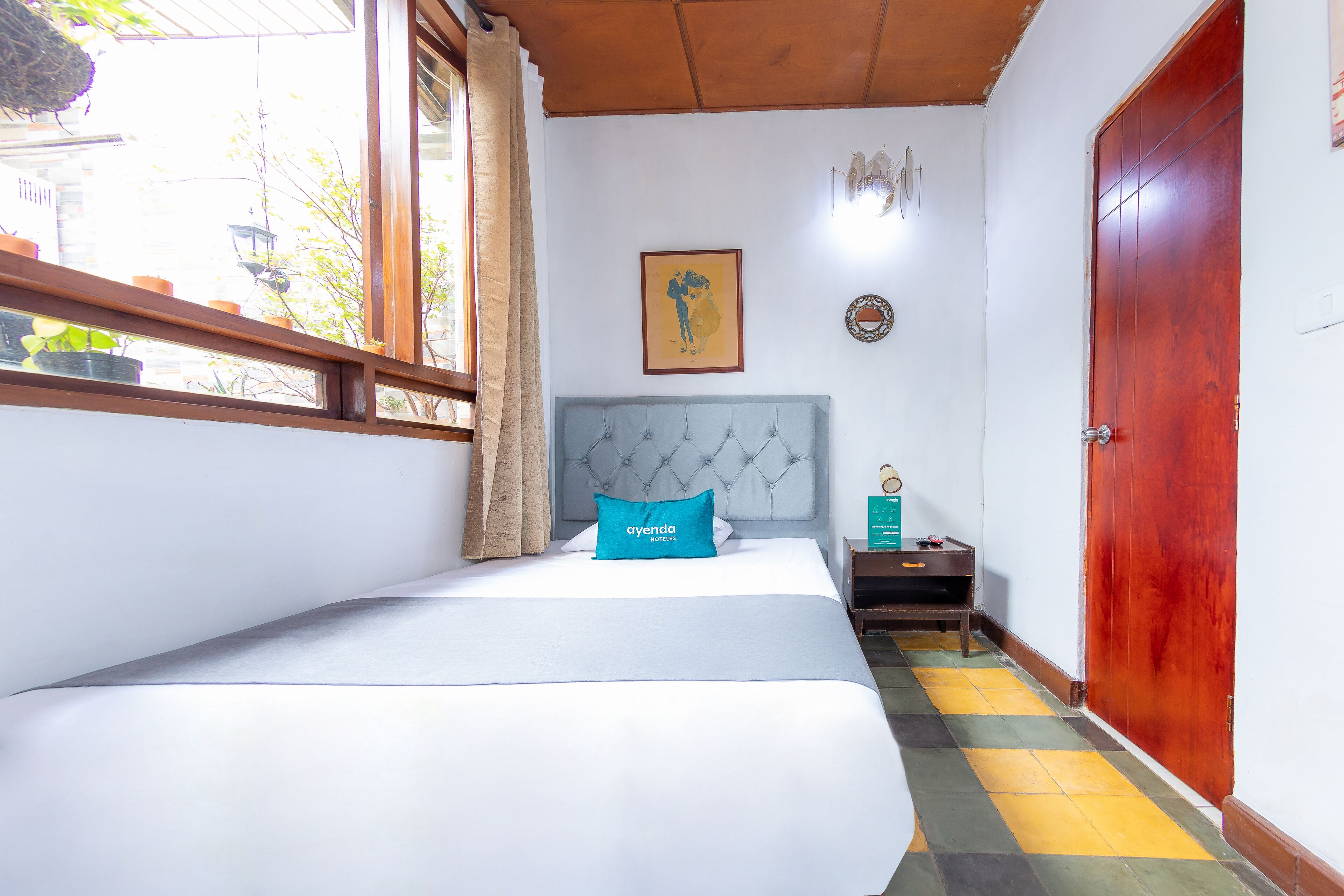A hotel room with a bed, a nightstand, a wooden door, and a window with curtains.