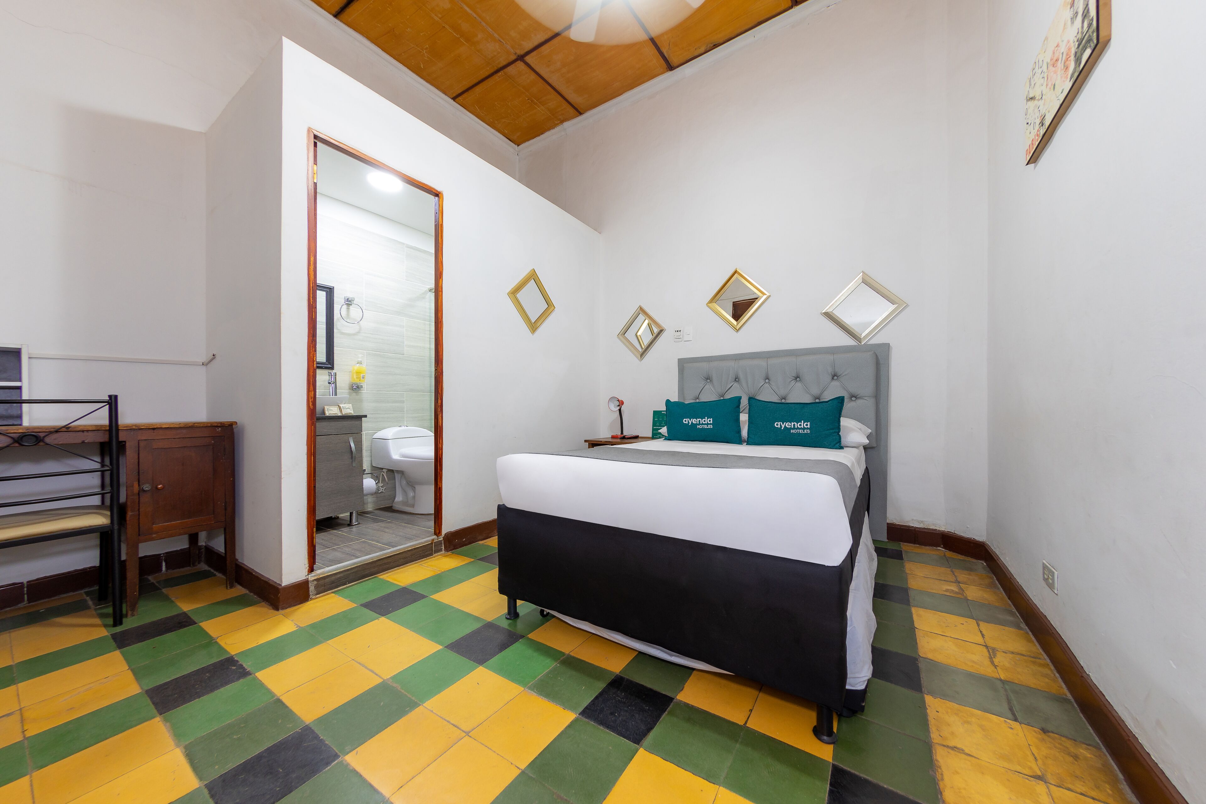Une chambre à coucher avec un lit, une armoire en bois, une salle de bain visible à travers une porte ouverte et un sol à carreaux.