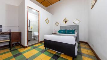 Une chambre à coucher avec un lit, une armoire en bois, une salle de bain visible à travers une porte ouverte et un sol à carreaux.