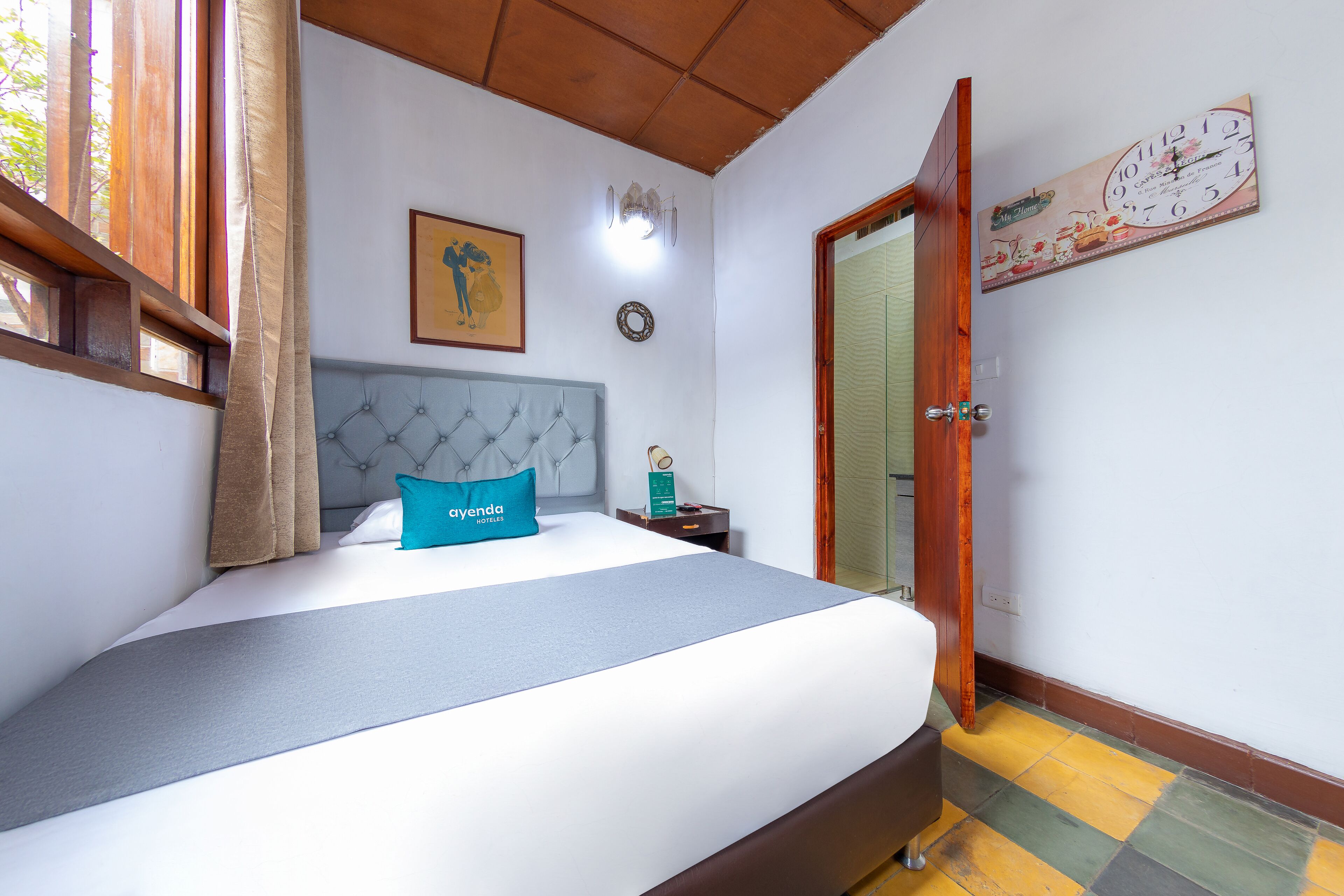 A hotel room with a bed, a nightstand, a wall clock, a framed picture, and a door leading to another room.