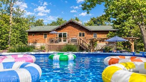 A wooden cabin with a swimming pool, surrounded by trees and a covered patio area.