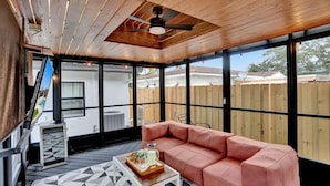 A covered patio with a wooden ceiling, a pink sofa, a coffee table with a tray, and a view of a backyard through large windows. - Casa De Amigos | Private Pool | Outdoor Oasis (Fort Lauderdale)