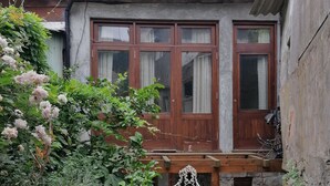 A courtyard with a stone wall, a wooden door, and a window with curtains. There are potted plants and a white chair. - Porto Old Downtown - Vérités Dame (Porto)