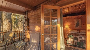 A wooden cabin with a porch, a bed with a quilt, and a window with curtains.