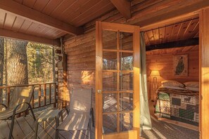 A wooden cabin with a porch, a bed with a quilt, and a window with curtains.
