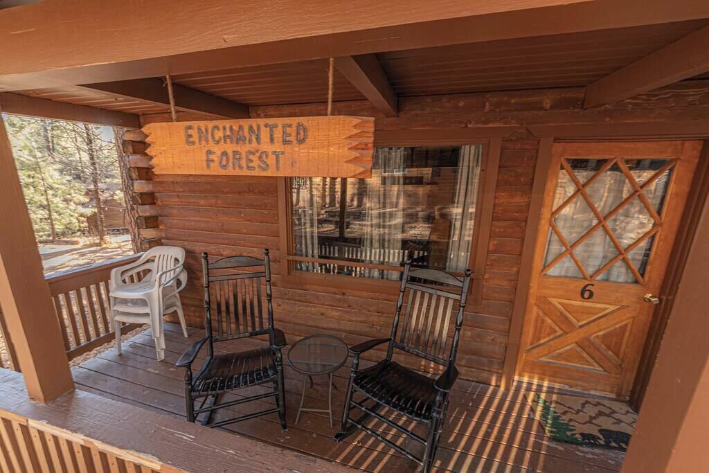 Eine Holzhütte mit einem Schild, auf dem "ENCHANTED FOREST" steht, einer Tür mit der Aufschrift "6" und zwei Stühlen sowie einem kleinen Tisch auf der Veranda.