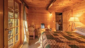 A cozy cabin room with a wooden interior, a bed with a quilt, a chair, a desk with a radio, and a door leading outside. - Forest Cabin 6 Enchanted Forest (Payson)