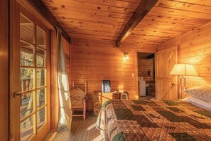 A cozy cabin room with a wooden interior, a bed with a quilt, a chair, a desk with a radio, and a door leading outside.