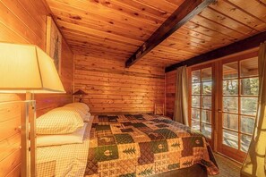 A wooden cabin bedroom with a quilted bedspread, a lamp, and a view through a glass door.
