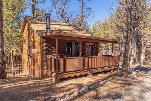 A rustic log cabin with a wooden deck, surrounded by trees.