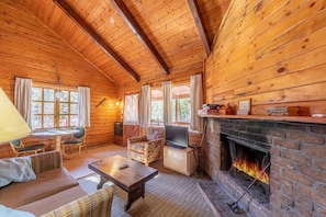 A cozy cabin interior with a fireplace, wooden walls, a sofa, a TV, and a dining table.