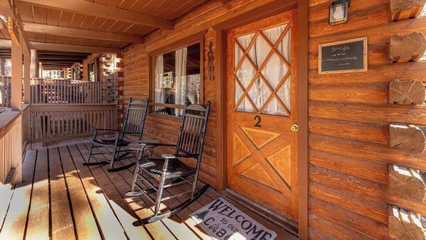A wooden cabin with a porch, two rocking chairs, a door with a number, and a welcome mat.