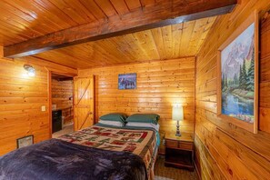 A wooden cabin bedroom with a bed, a nightstand, a painting, and a door leading to another room. - Forest Cabin 1 Hunters Blind (Payson)