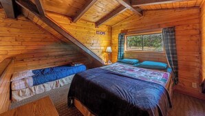A cozy cabin room with a loft bed, a smaller bed, and a window with curtains. - Forest Cabin 1 Hunters Blind (Payson)