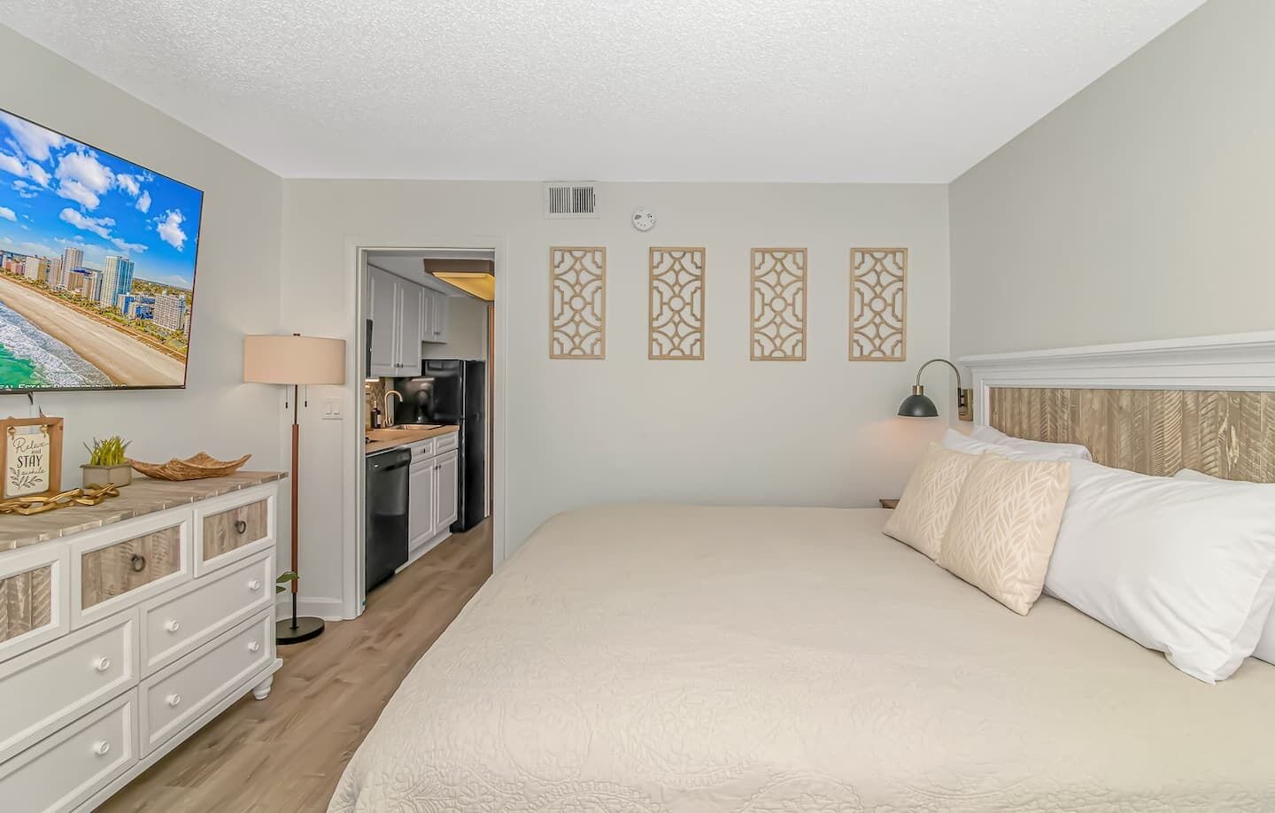 A hotel room with a large bed, a desk with a lamp, a TV on the wall, and a kitchenette.