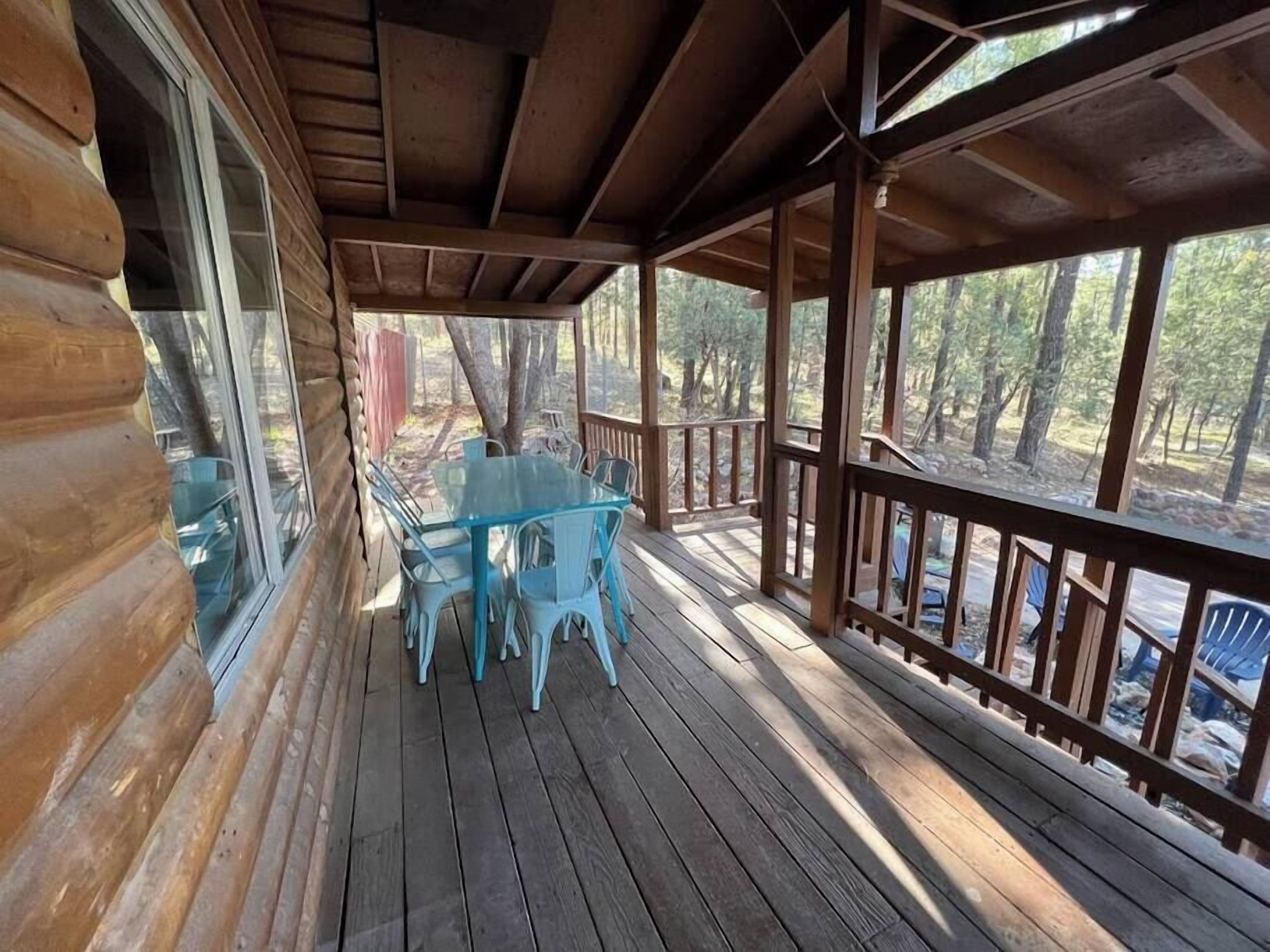 A covered wooden deck with a glass table and chairs, surrounded by a wooden railing and trees.