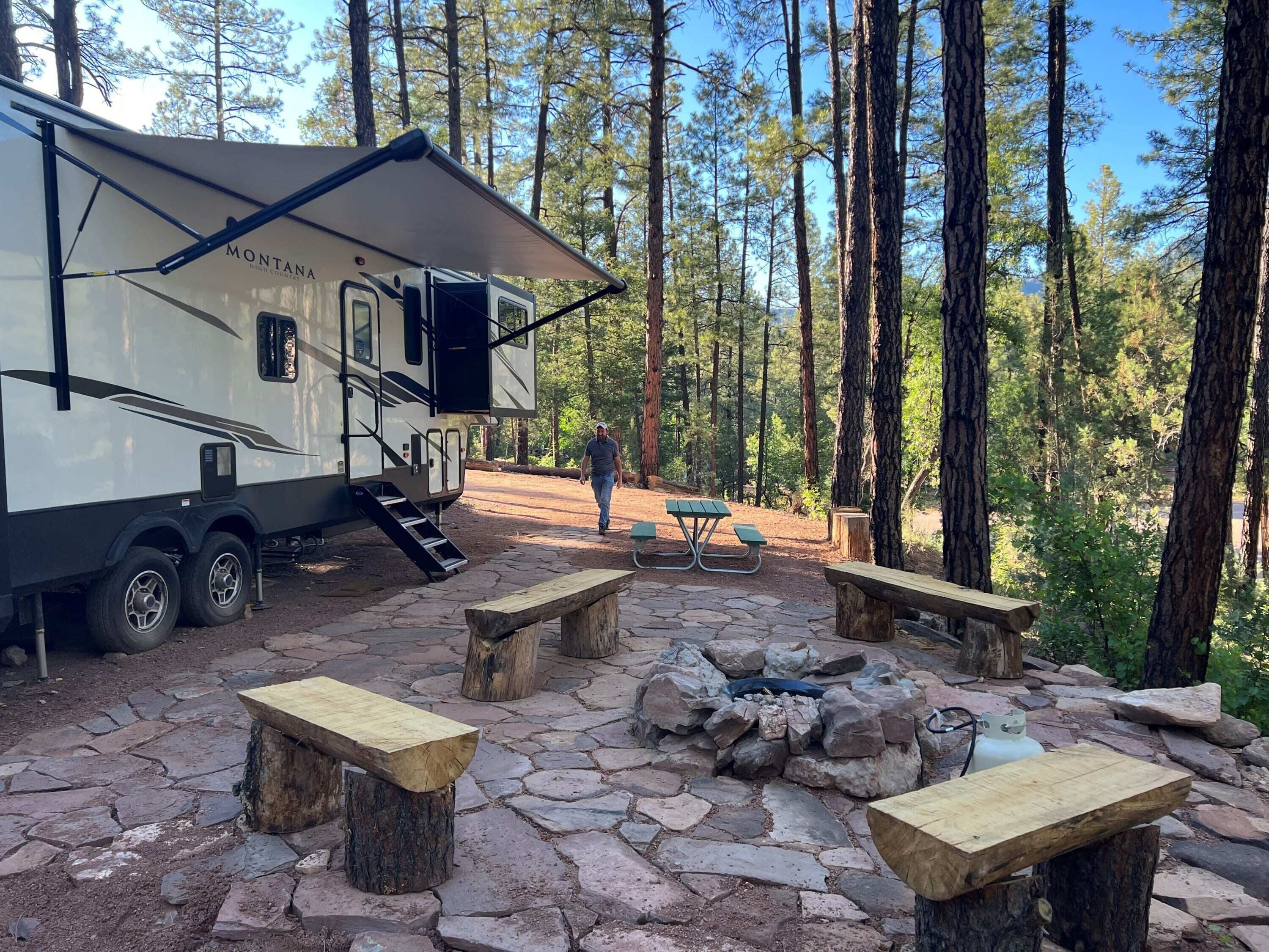 A campsite with a large RV, picnic tables, and a fire pit surrounded by trees.