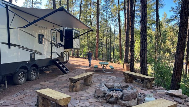 Un camping con una caravana grande, mesas de picnic y un pozo para fuego rodeado de árboles.