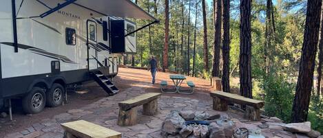 A campsite with a large RV, picnic tables, and a fire pit surrounded by trees.