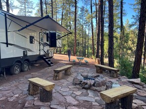 A campsite with a large RV, picnic tables, and a fire pit surrounded by trees.