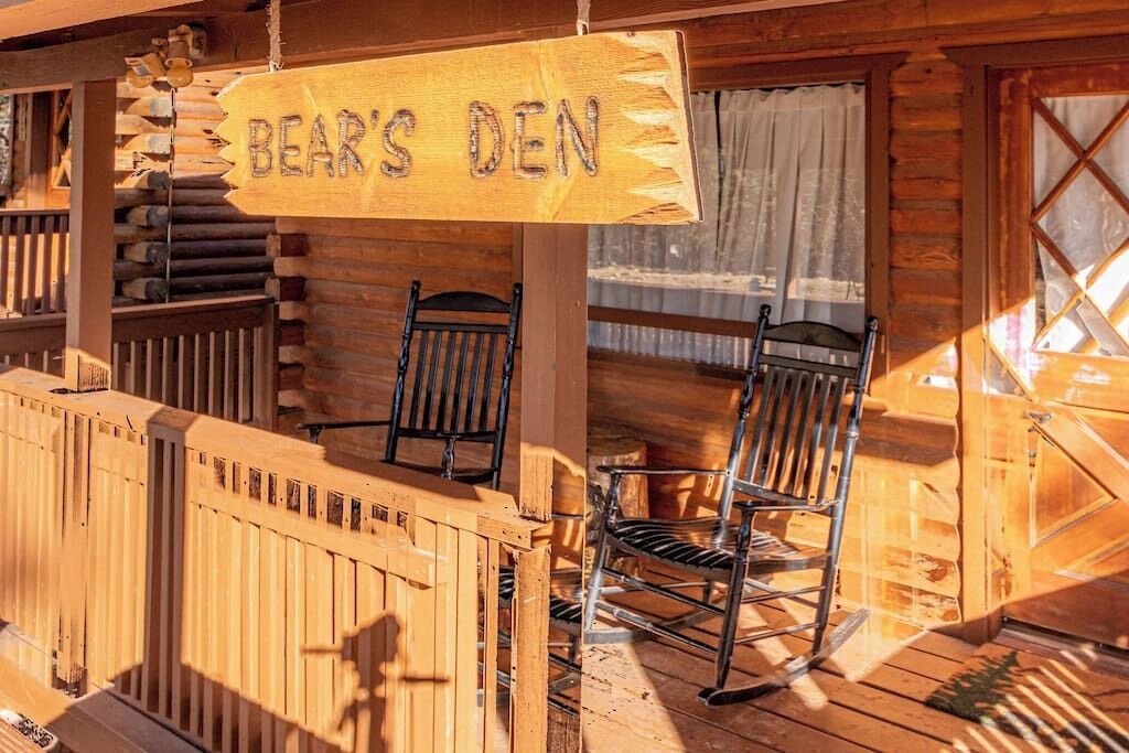 A wooden cabin with a sign reading "BEAR'S DEN" and two rocking chairs on the porch.