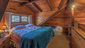 A cozy attic bedroom with a wooden ceiling, a bed with a quilt, a nightstand, a dresser, and a window with curtains. - Forest Cabin 3 Bear's Den (Payson)