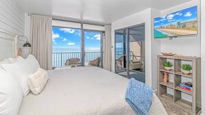 A bedroom with a large bed, a TV, a bookshelf, and a balcony with a view of the sea.