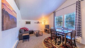 Sala de estar con sofá, mesa de centro, mueble para televisor y mesa de comedor con sillas. Hay dos ventanas con cortinas, un cuadro en la pared y una alfombra en el suelo.