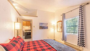Een slaapkamer met een bed, een raam met geruit gordijnen, een schilderij aan de muur en een kitchenette op de achtergrond.