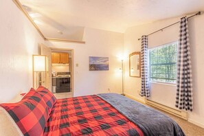 A bedroom with a bed, a window with checkered curtains, a painting on the wall, and a kitchenette in the background. - Quaint condo retreat 12 Ridgeview Condo (Payson)