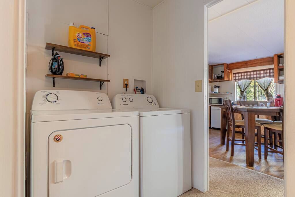 A laundry area with two front-loading washers, shelves with cleaning supplies, and a view into a dining area with wooden furniture.