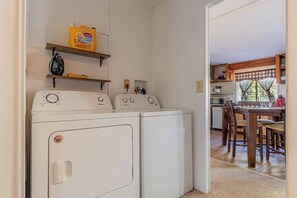 A laundry area with two front-loading washers, shelves with cleaning supplies, and a view into a dining area with wooden furniture. - Forest Home Retreat 11 Ridgeview Retreat (Payson)
